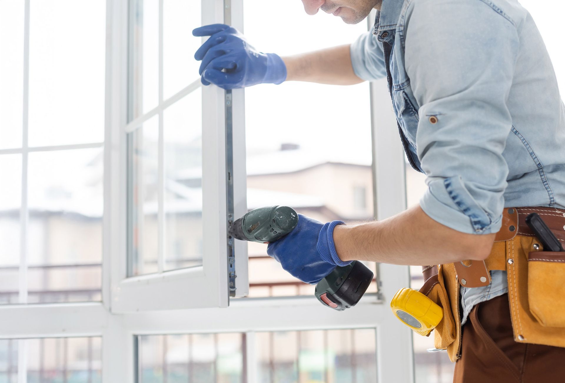 Man in blue gloves using a power drill to install a white window.