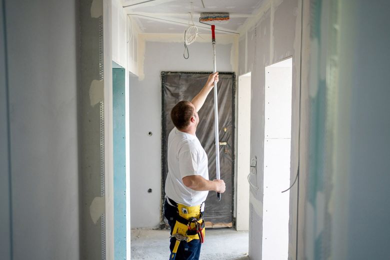 A man in a white shirt and tool belt uses a roller to paint a ceiling in a hallway under construction.