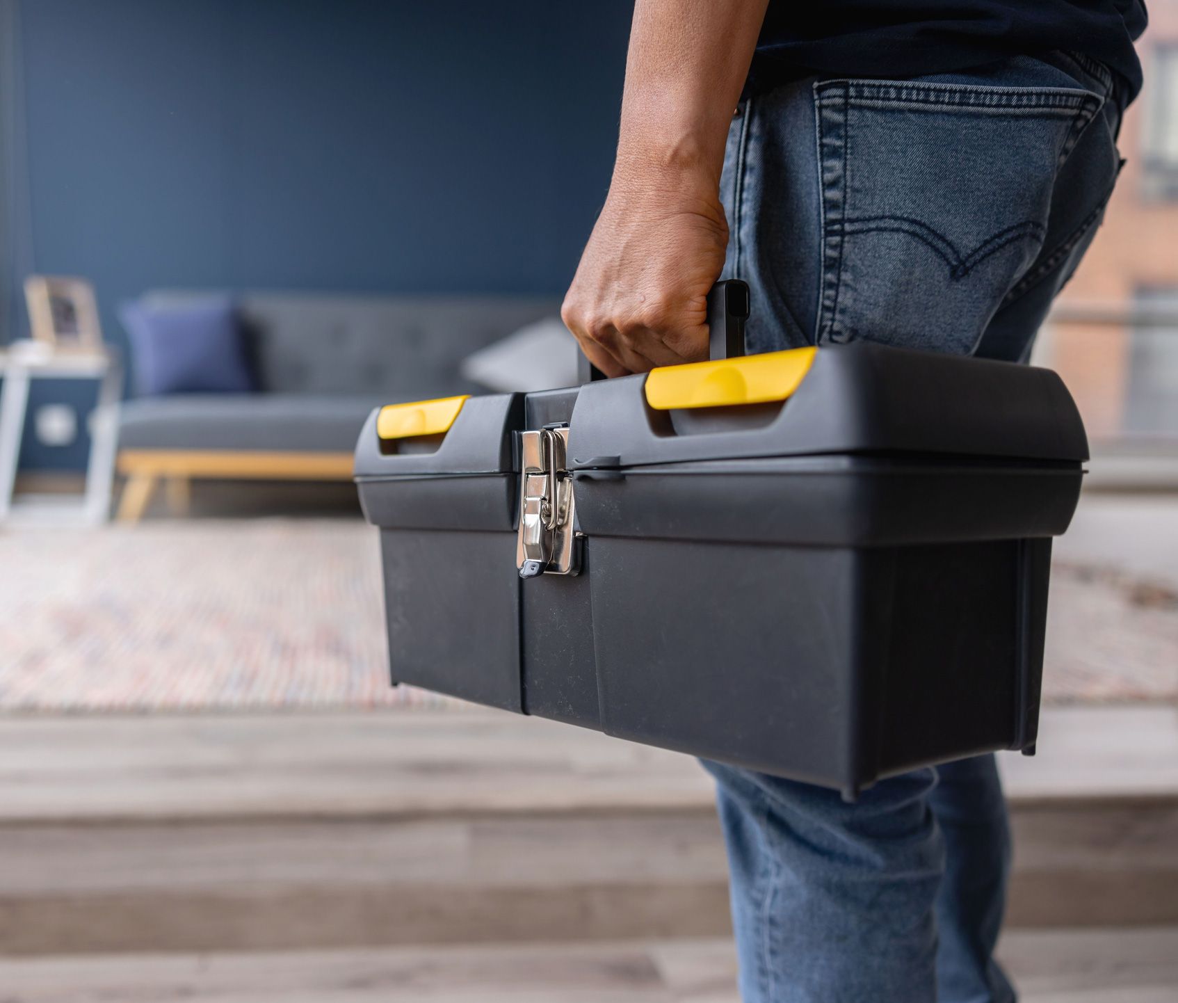 Person carrying a black toolbox with yellow accents indoors, near a couch.