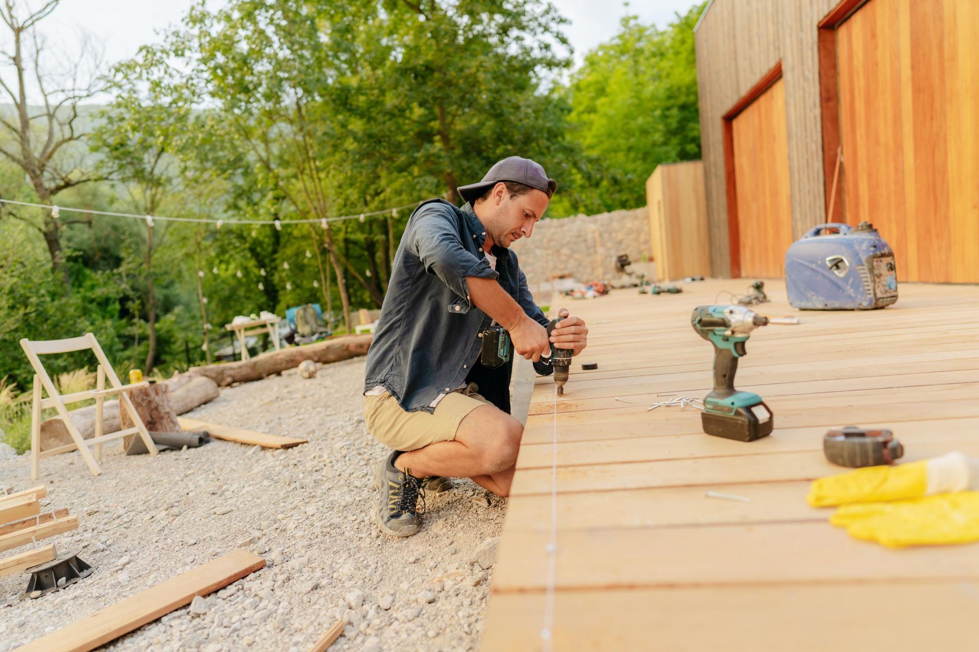 Man in shorts using a drill to build a wooden deck outdoors; sunny day.