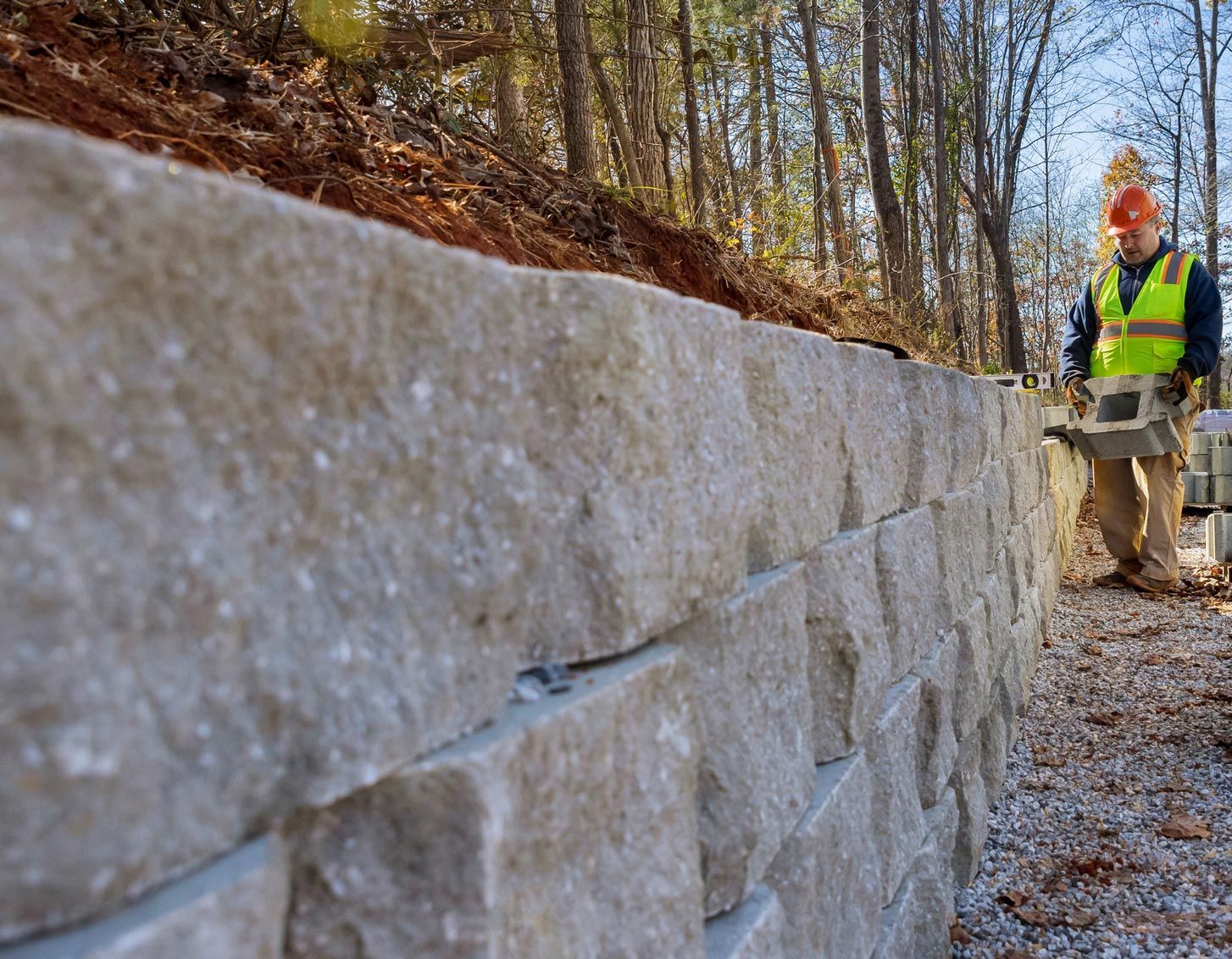 Man constructing a retaining wall with light gray stone blocks in a wooded area.