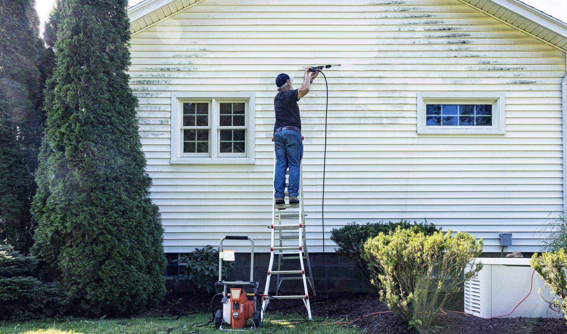 Person on a ladder power washing a white house with green algae.