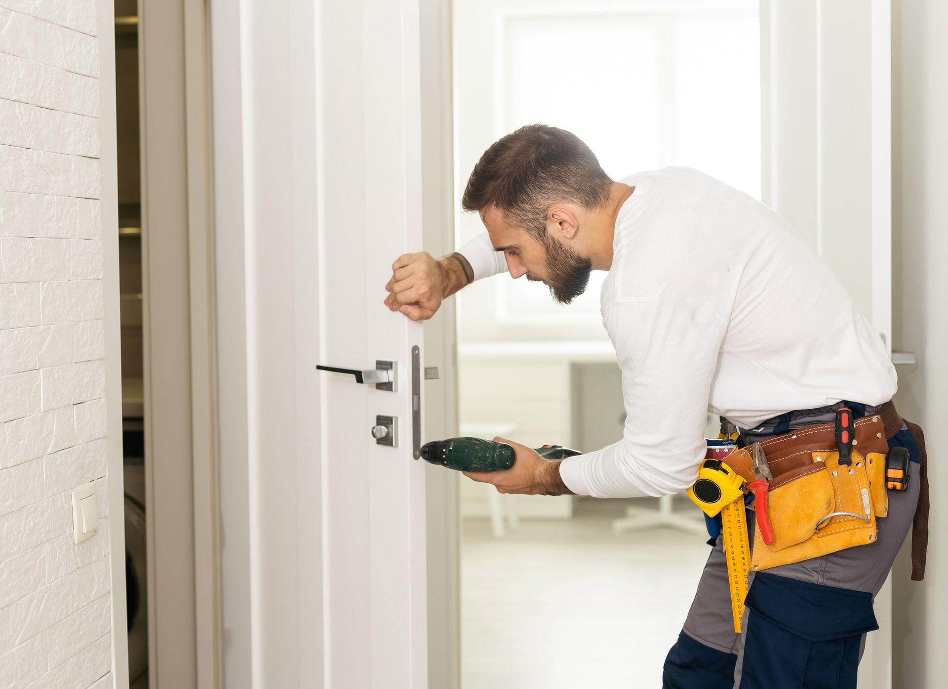 A handyman installing a doorknob with a drill in a white doorway.