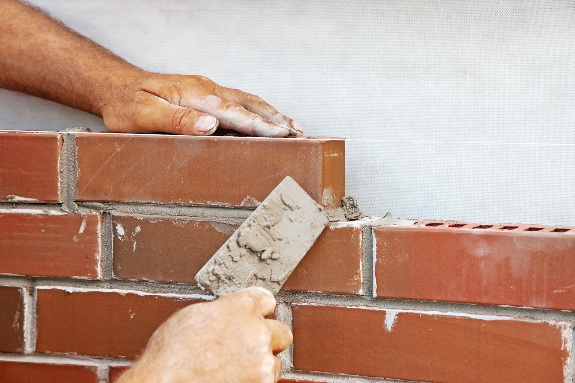 Hands of a bricklayer placing a brick with mortar, against a white wall.