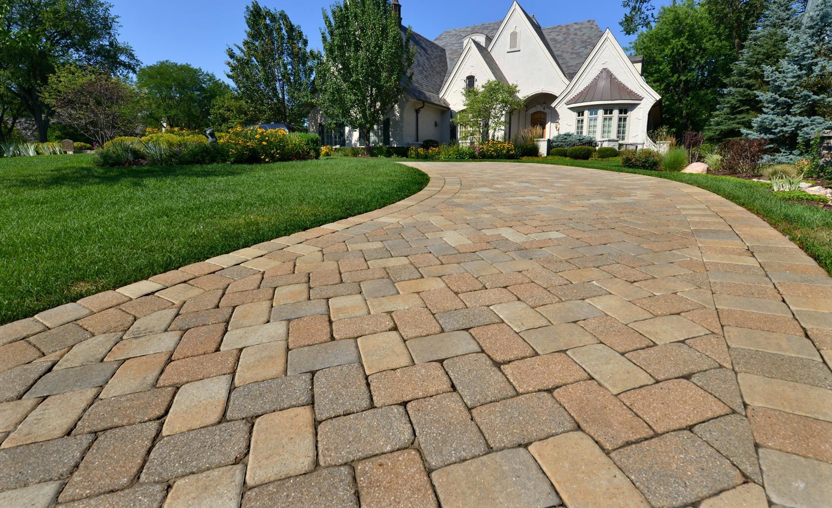 Brick driveway leading to a light-colored house with landscaping on a sunny day.