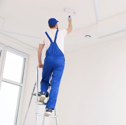 A painter in blue overalls and hat painting a white ceiling while standing on a stepladder.
