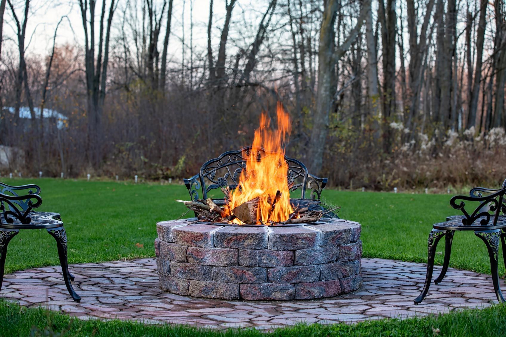 Fire pit with burning flames in a backyard, surrounded by brick pavers, two benches, and trees.