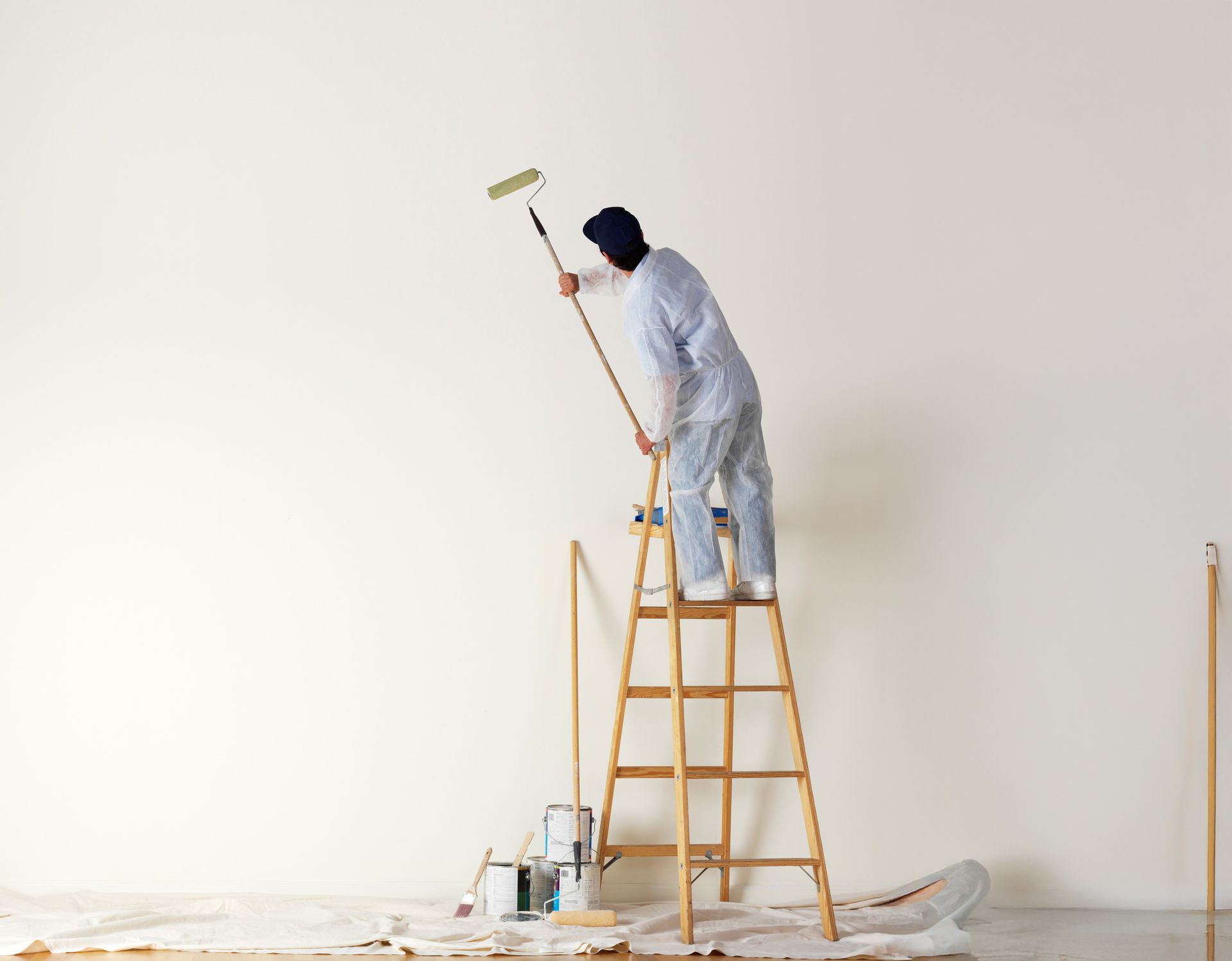 Person in a coverall painting a white wall with a roller while standing on a wooden ladder.
