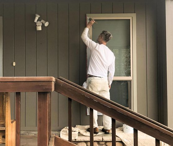 Person painting window trim on a house exterior, reaching up with brush.