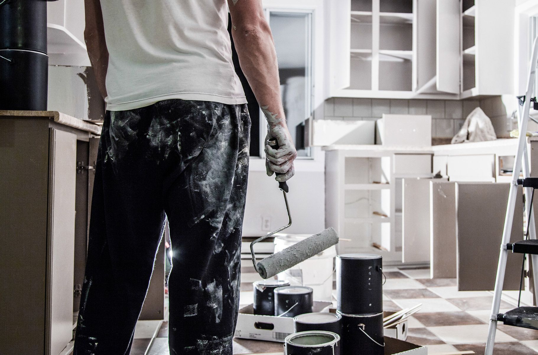 Person painting kitchen cabinets, wearing paint-splattered pants and gloves, holding a paint roller.