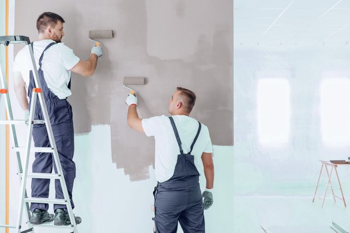 Two painters in overalls apply gray paint to a wall using rollers. One is on a ladder.