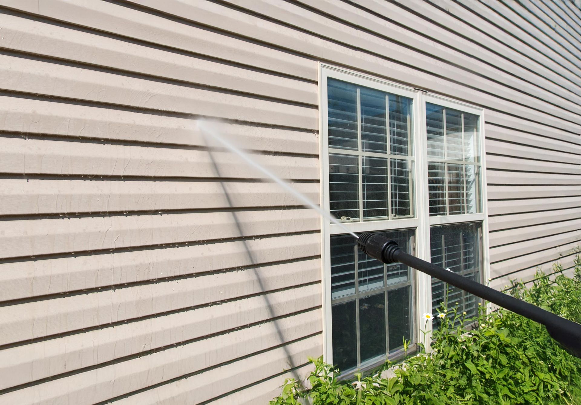 Power washing tan vinyl siding near a window with white trim.