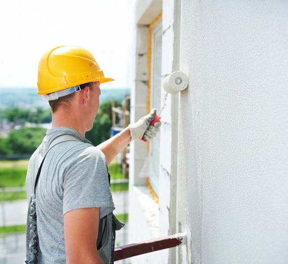 Construction worker in a yellow hard hat and gray overalls painting exterior wall with roller.
