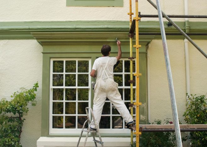 Painter on a ladder, painting green trim around a window on a white house exterior.