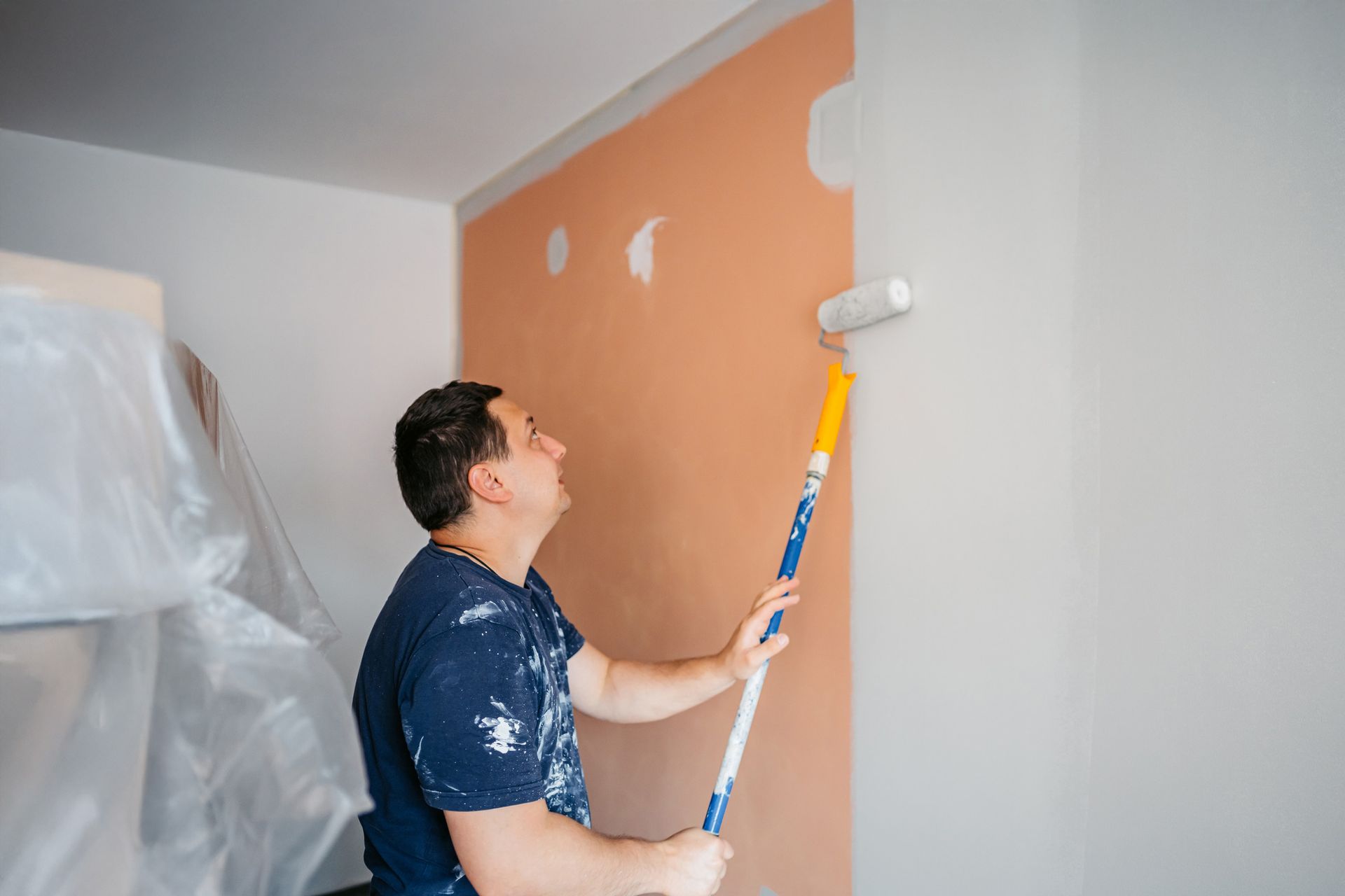 Man painting a wall with a roller, using a paint roller on a long pole.
