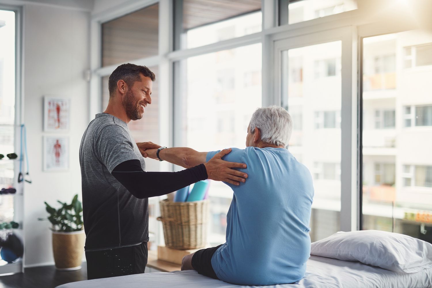 A physical therapist assists an elderly man with arm exercises in a bright clinic. Both are smiling.