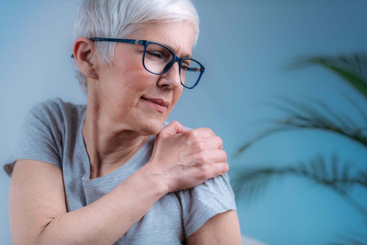 Woman with grey hair, glasses, in pain, touching her shoulder; light blue background.