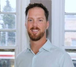 A man with a beard smiles in front of a window, wearing a light blue button-down shirt.