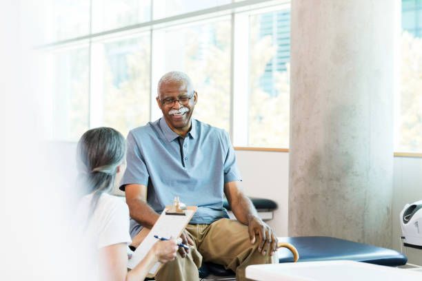 An older Black man smiles while conversing with a healthcare provider holding a clipboard in a light-filled clinic.