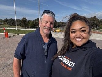 Man and woman smiling for selfie outdoors; the woman wears a dark hoodie.