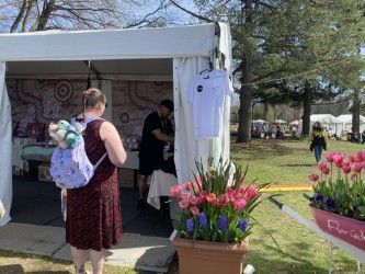 Person looking at a vendor booth with a shirt hanging outside. Flowers and people in the background on a sunny day.