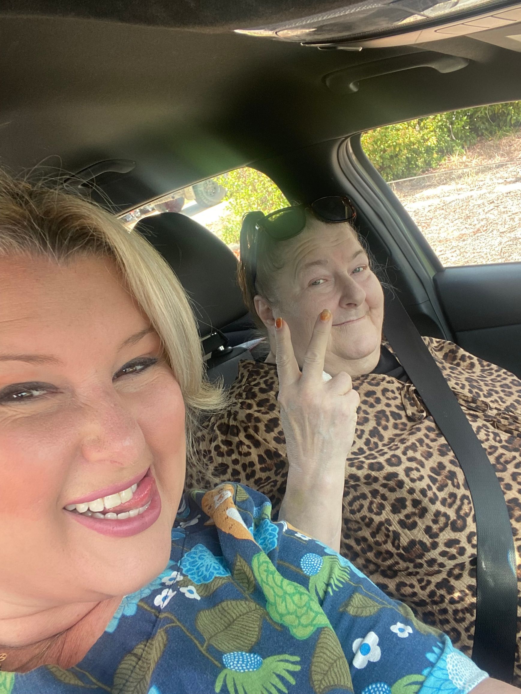 Woman smiling, taking a selfie with a man wearing sunglasses outdoors on a sunny day.