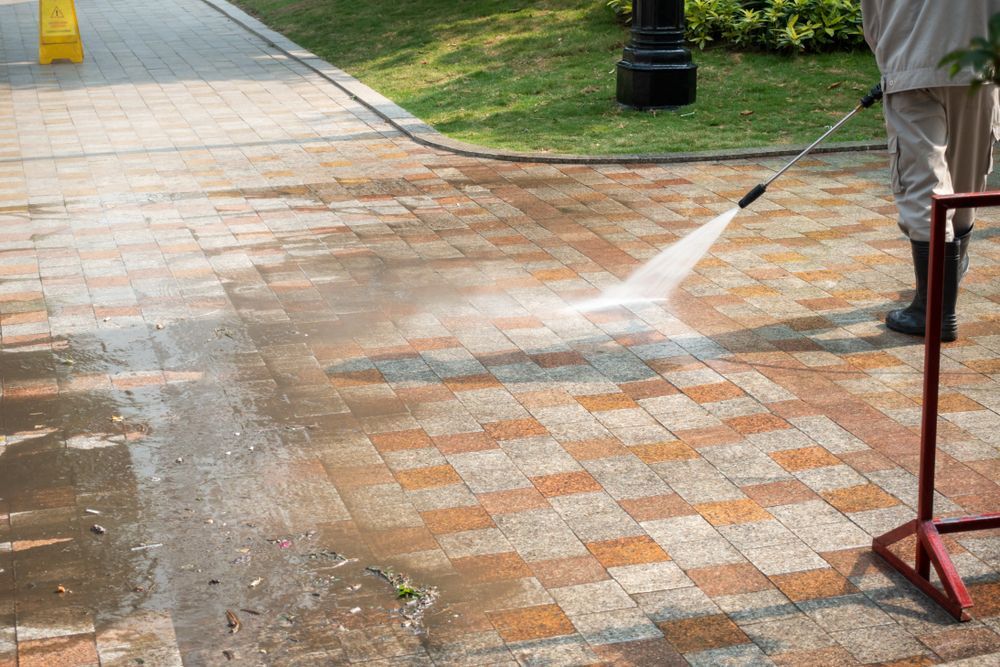Person pressure-washing a brick walkway outdoors with water spraying across the pavement