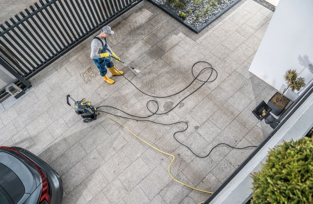 Worker power-washing a driveway with hoses and equipment beside a parked car and gate