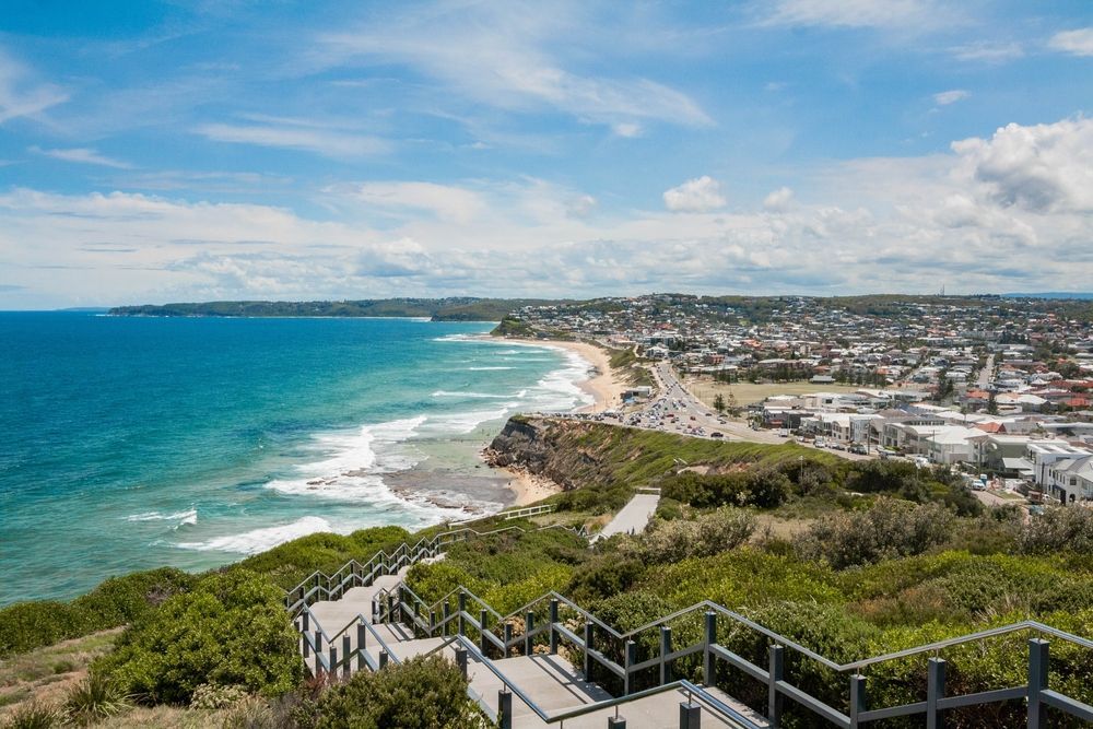 Coastal cityscape with turquoise ocean, sandy beach, and hillside walkway overlooking the shoreline