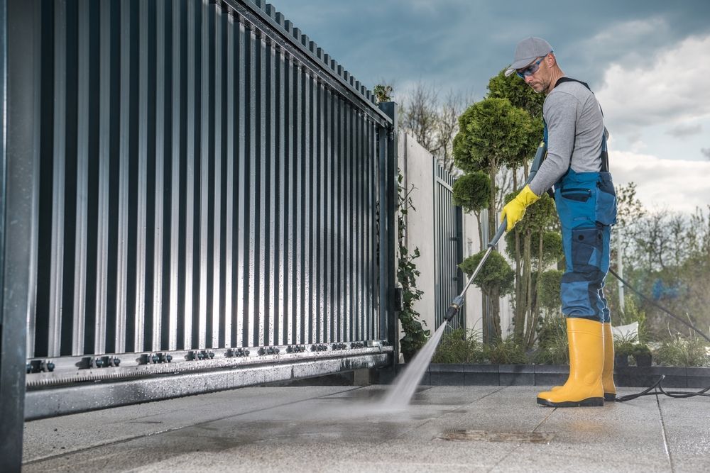 Worker pressure-washing a driveway beside a black metal fence, wearing yellow boots and gloves.
