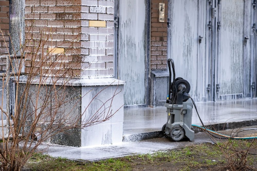 Yellow-and-black pressure washer on a wet patio, spraying water near stacked firewood and a grass edge