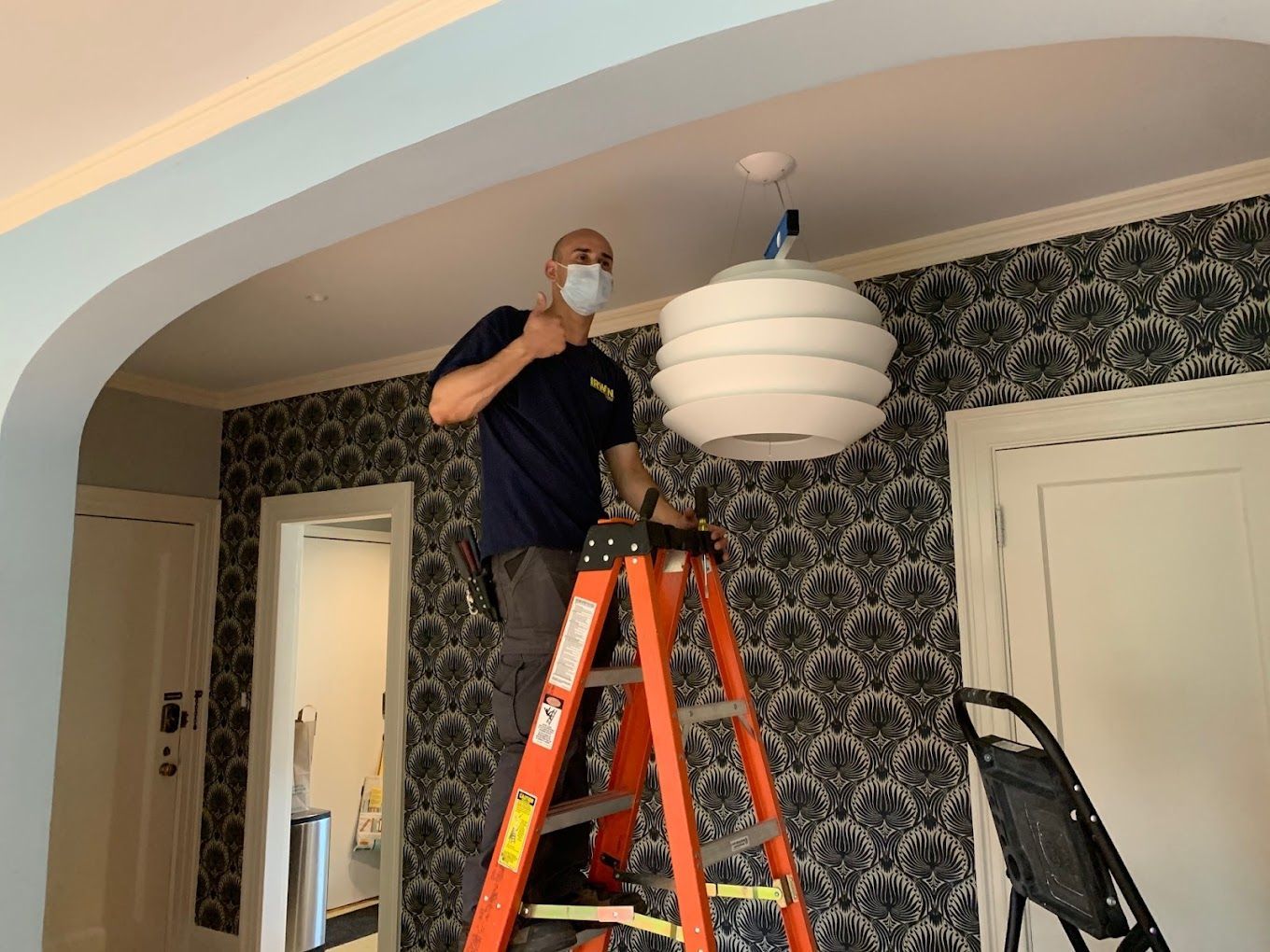 A man wearing a mask is standing on a ladder fixing a light fixture.