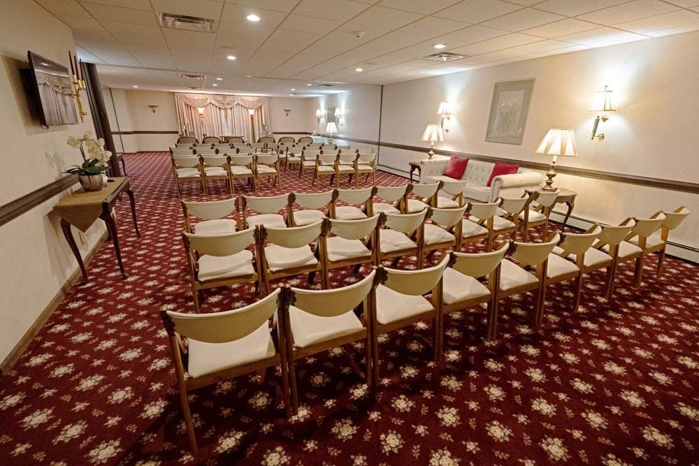Empty room with chairs arranged for a ceremony; red patterned carpet; white walls; lamps and artwork.