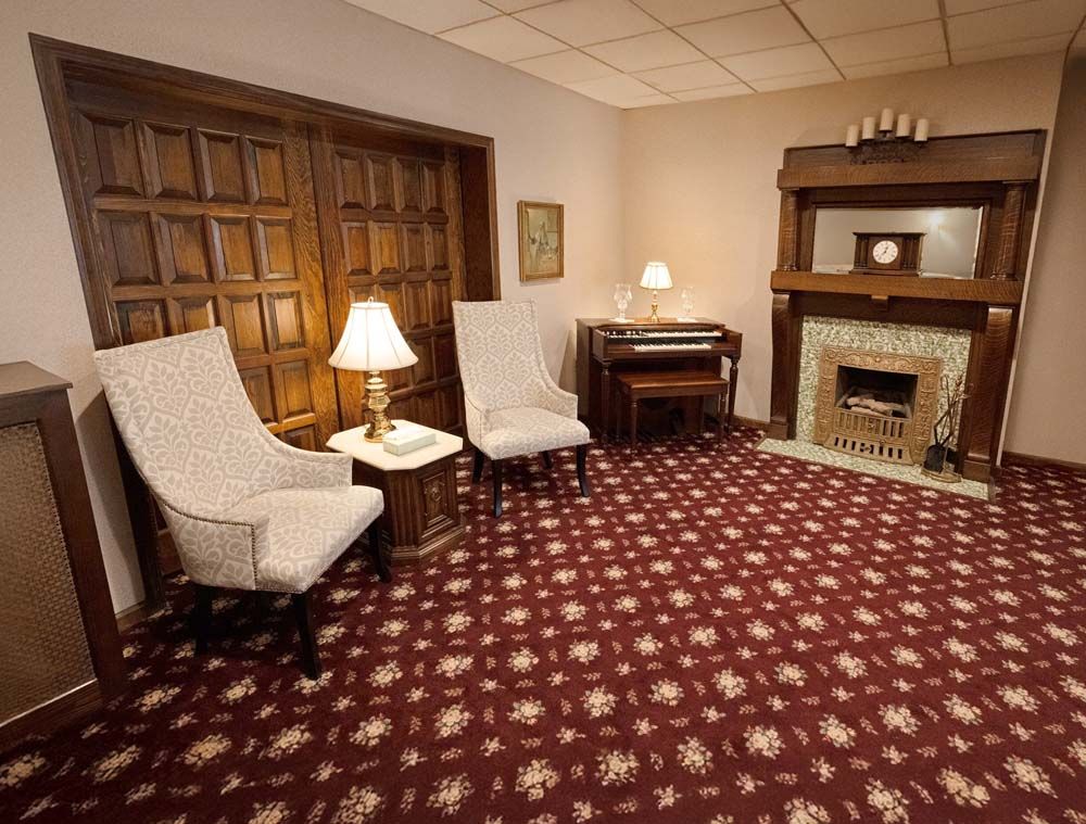 A warmly lit room with armchairs, a piano, fireplace, and patterned red carpet.