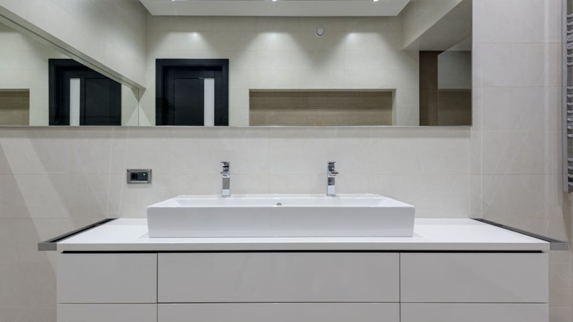 Un moderno mueble de baño con un lavabo blanco alargado, dos grifos plateados, un espejo grande 