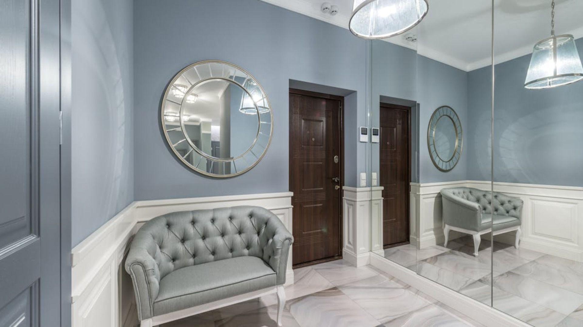 Un recibidor con paredes de color azul polvoriento, revestimiento de madera blanca, un sofá capitoné, un espejo redondo 