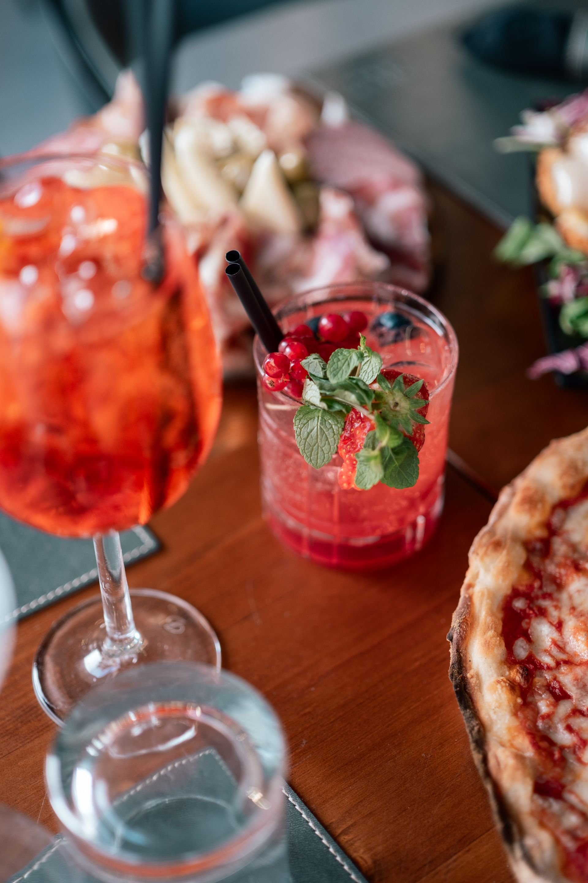 Bicchieri da cocktail e una pizza su un tavolo di legno, con bevande guarnite con menta e frutta.