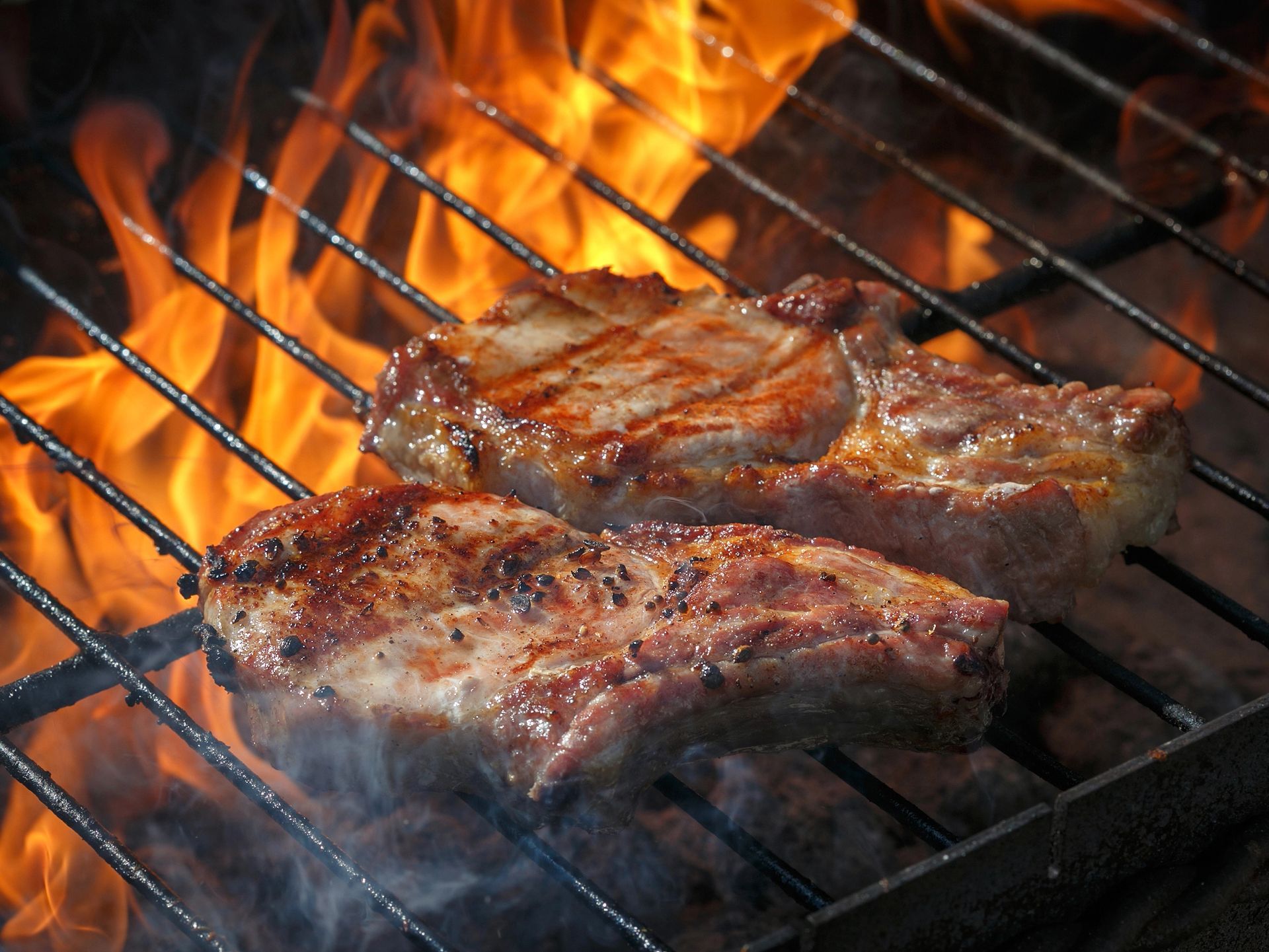 Bistecche grigliate su fiamme libere su un barbecue, con segni di grigliatura bruciacchiati e fumo che sale.