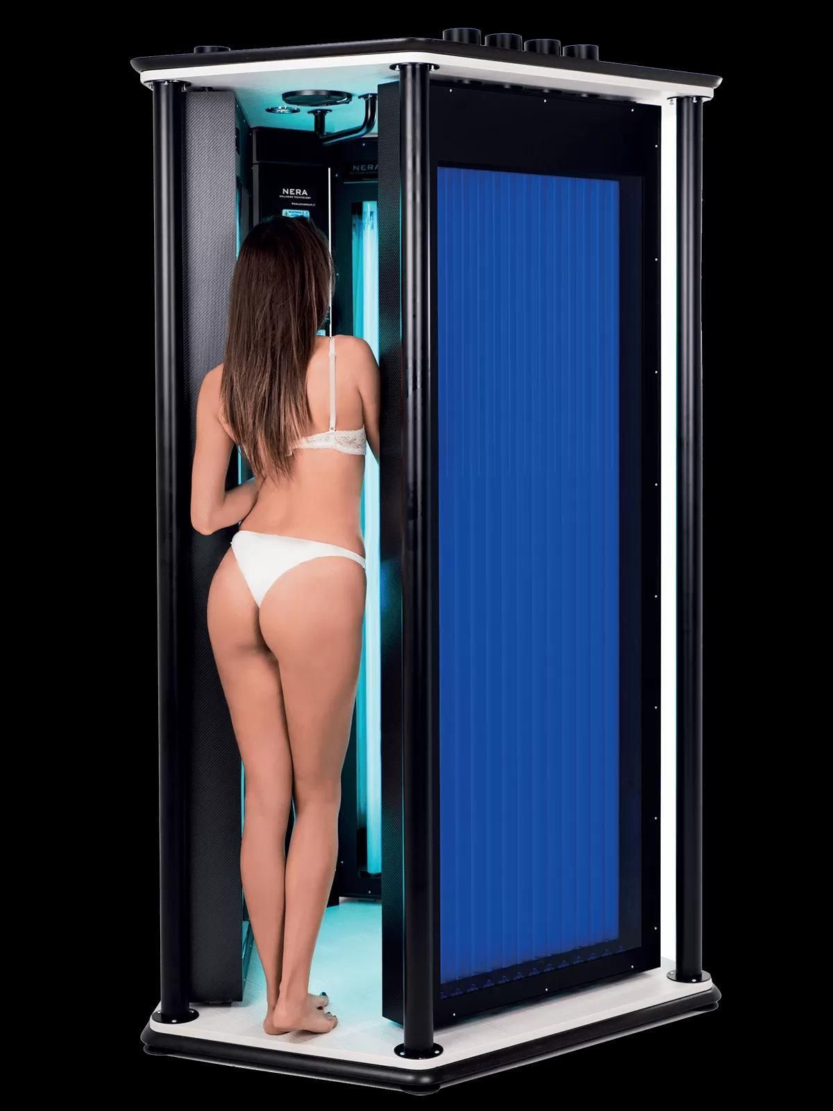 Una donna in bikini bianco è in piedi in una cabina abbronzante con un pannello luminoso blu.