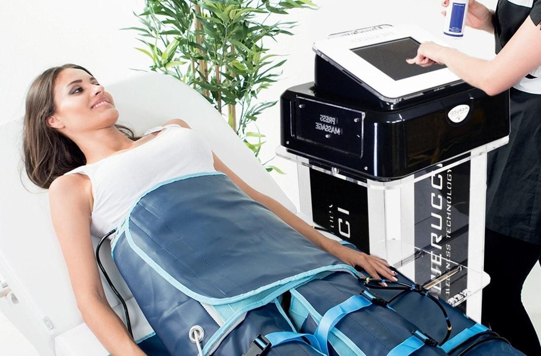 Donna sottoposta a terapia di compressione alle gambe in una spa. Fasciatura blu, macchina nera, presenza di un tecnico.