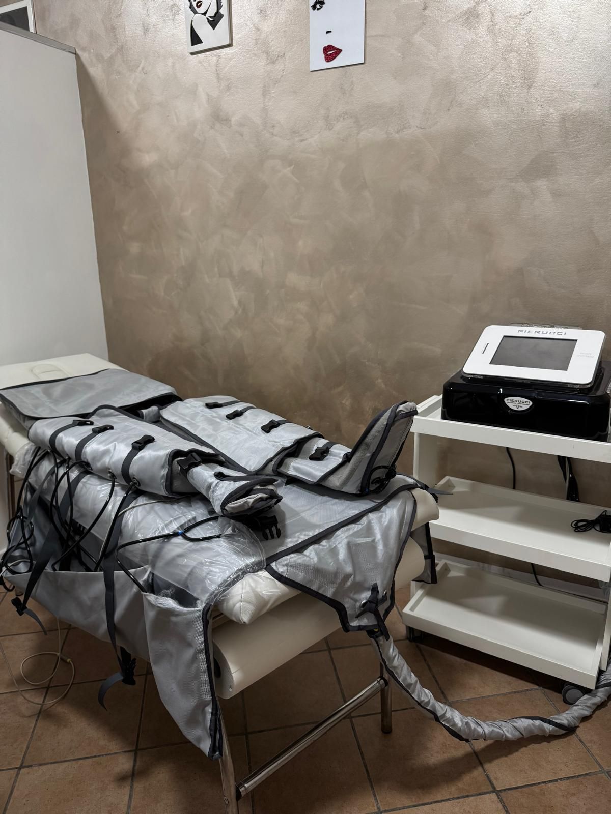 Persona sdraiata su un lettino da massaggio con dispositivo per terapia compressiva. Toni beige e argento. Carrello bianco a lato.