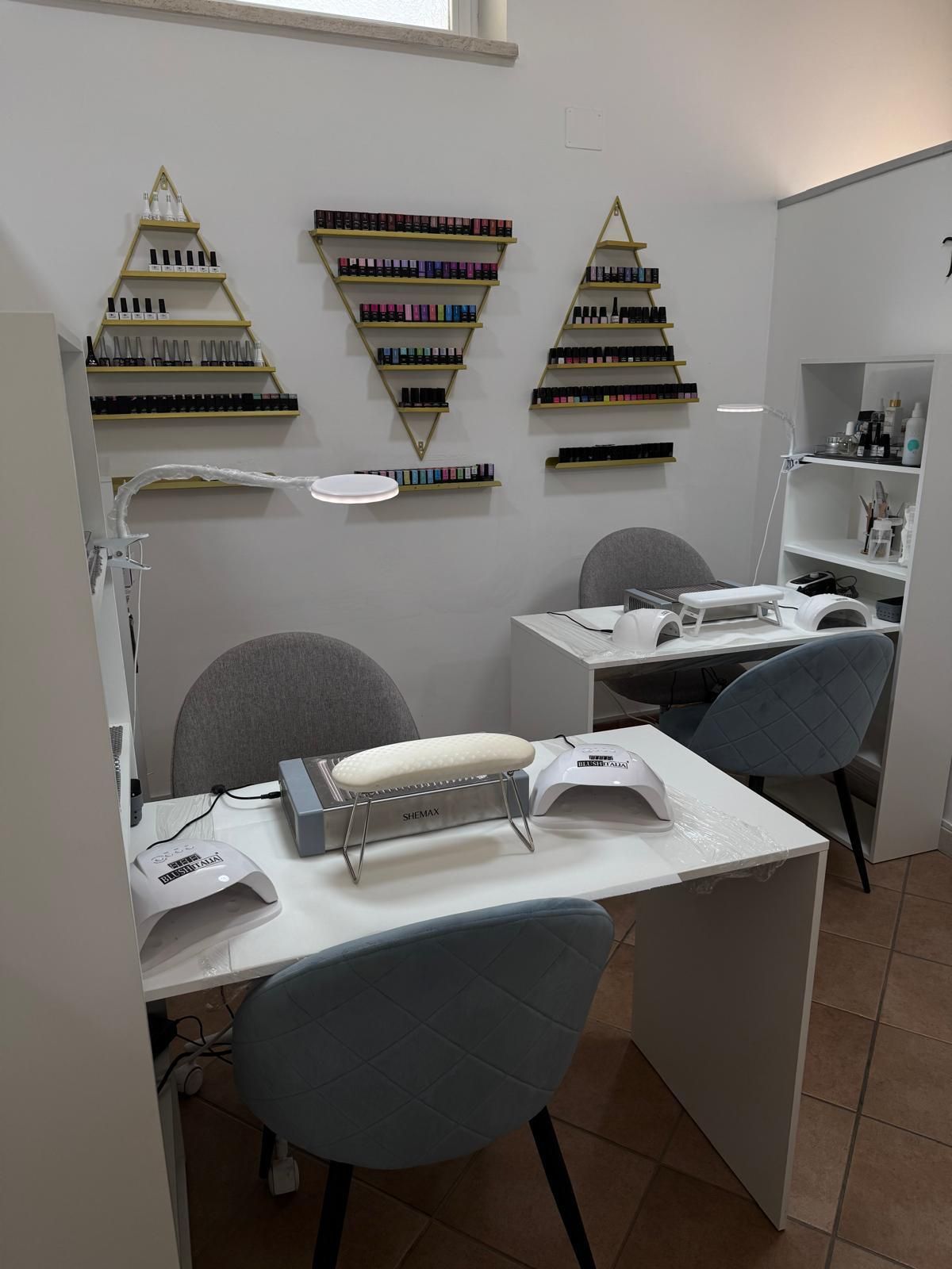 Interno di un salone per unghie con tavoli bianchi, sedie blu e scaffali con smalti.