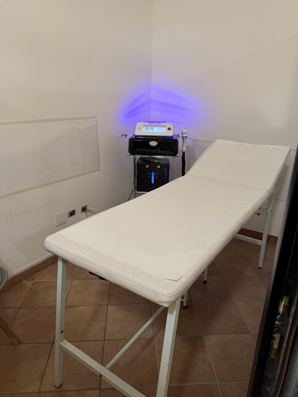 Sala visite con lettino bianco, dispositivo medico con luce blu e pavimento piastrellato.