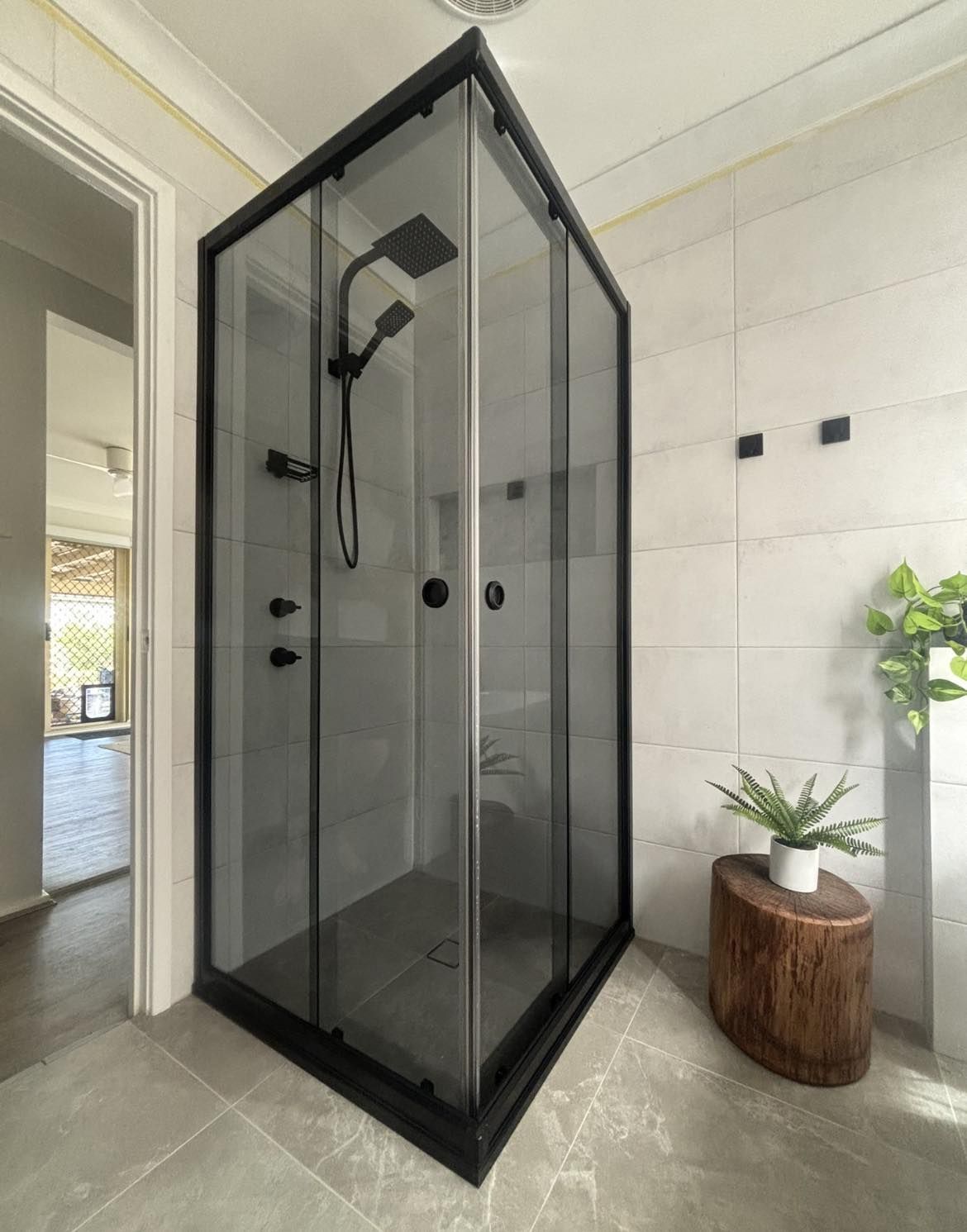 Black-framed, glass shower enclosure in modern bathroom. Shower fixtures are black, and a small plant sits on a wood stool — Cessnock Glass in Cessnock, NSW