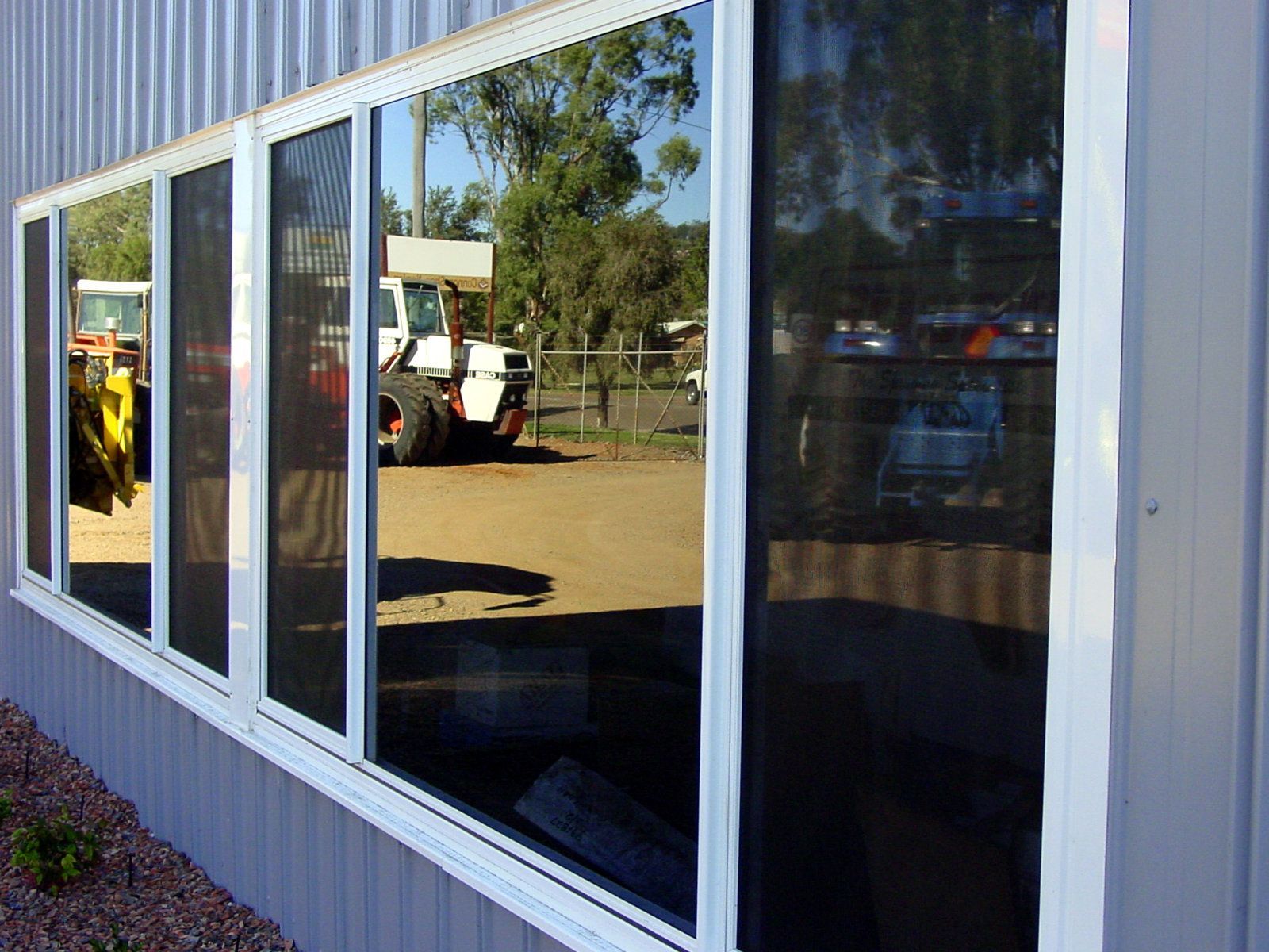 Long row of windows with reflective black glass on a gray building.  A tractor and trees are reflected in the glass — Cessnock Glass in Cessnock, NSW