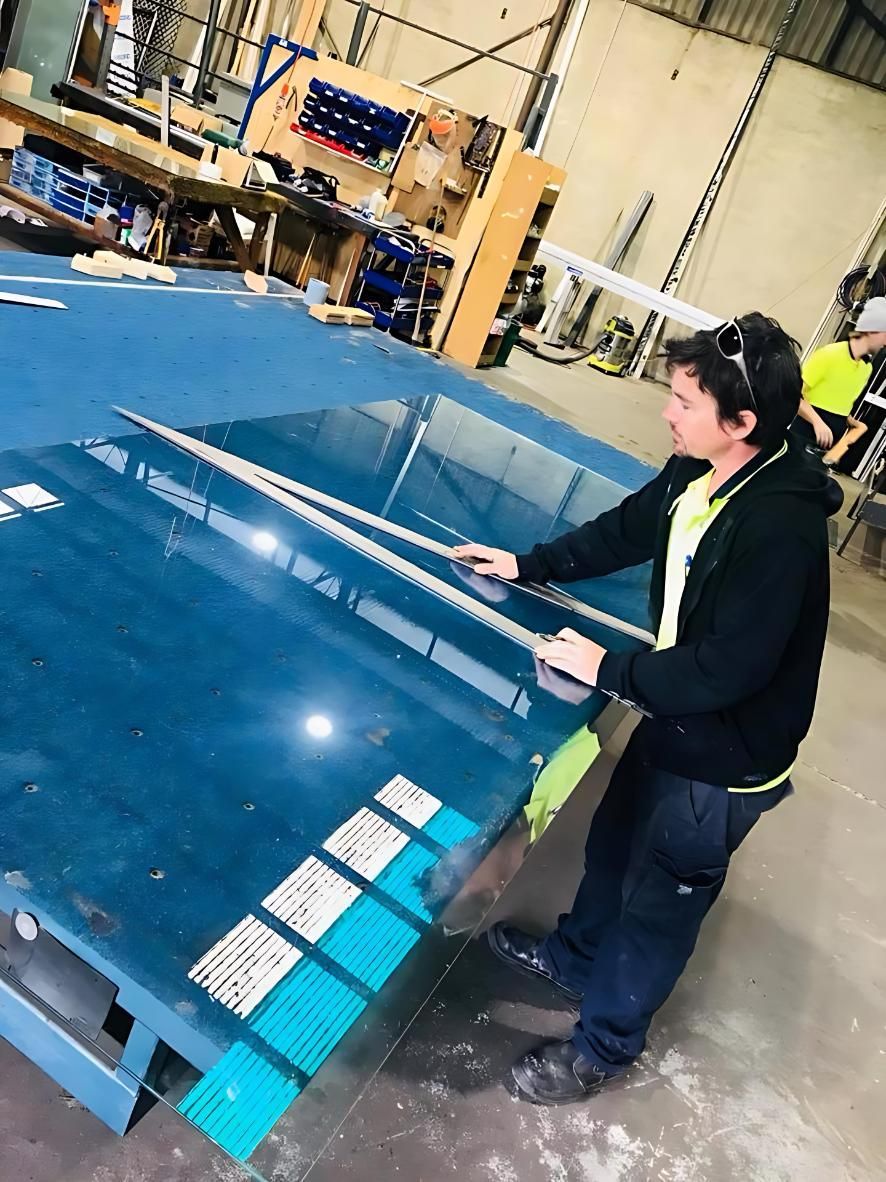 Worker measuring and cutting a large glass sheet on a blue workbench inside an industrial workshop.