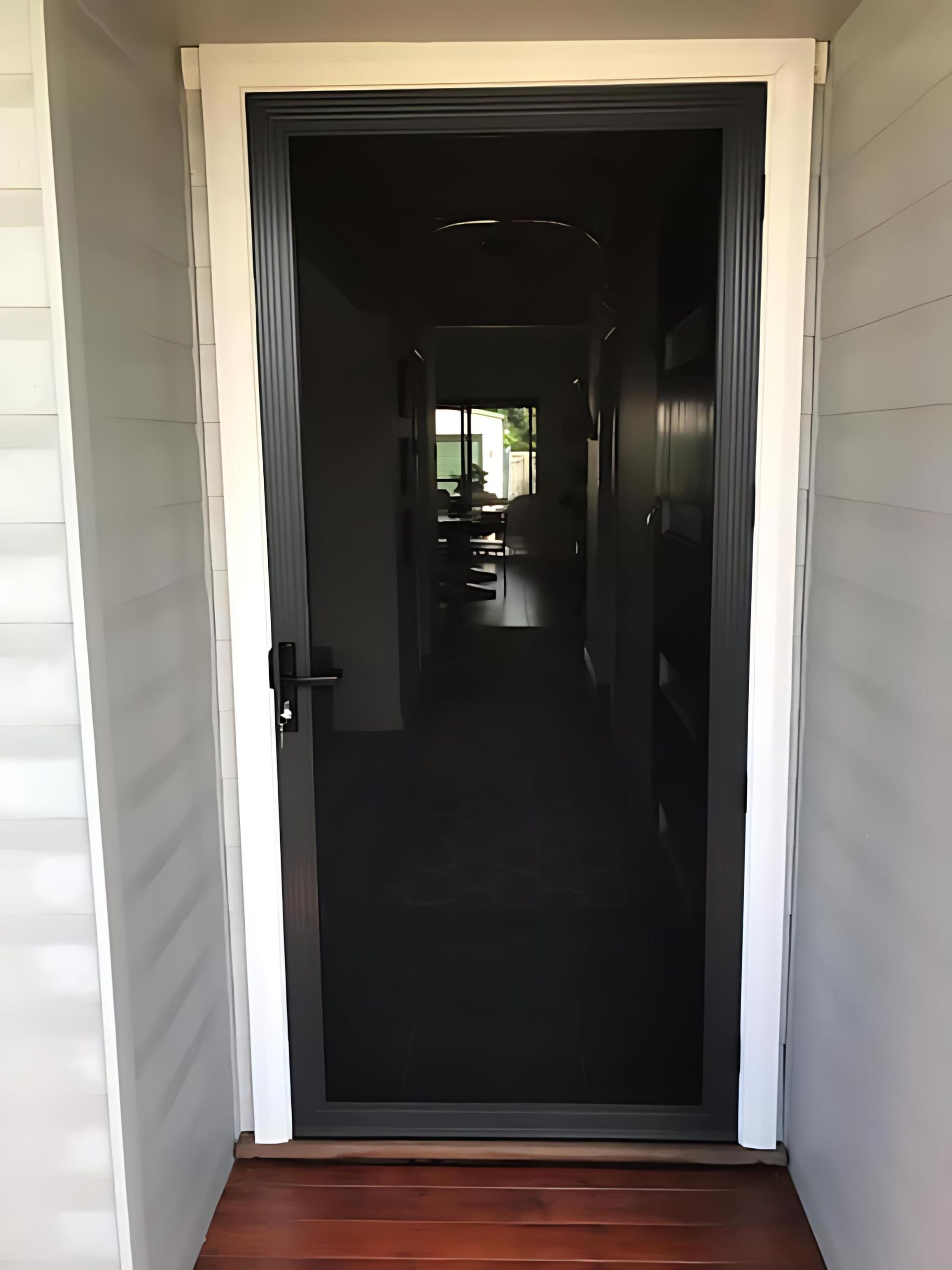 A black security door in a white-framed doorway, leading to a dimly lit hallway with wooden flooring — Cessnock Glass in Cessnock, NSW