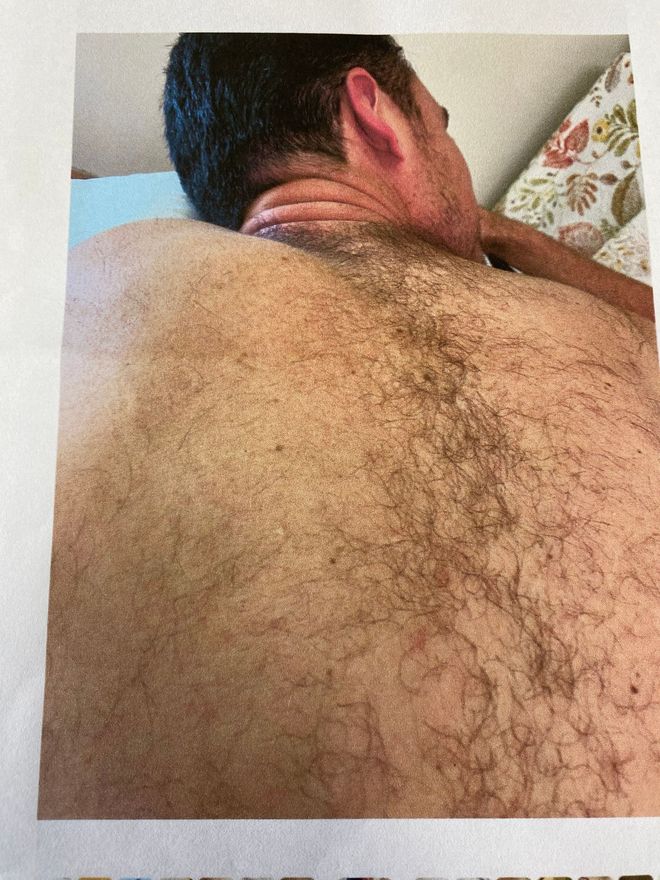 Man's hairy back and neck, viewed from behind. He is on a bed.