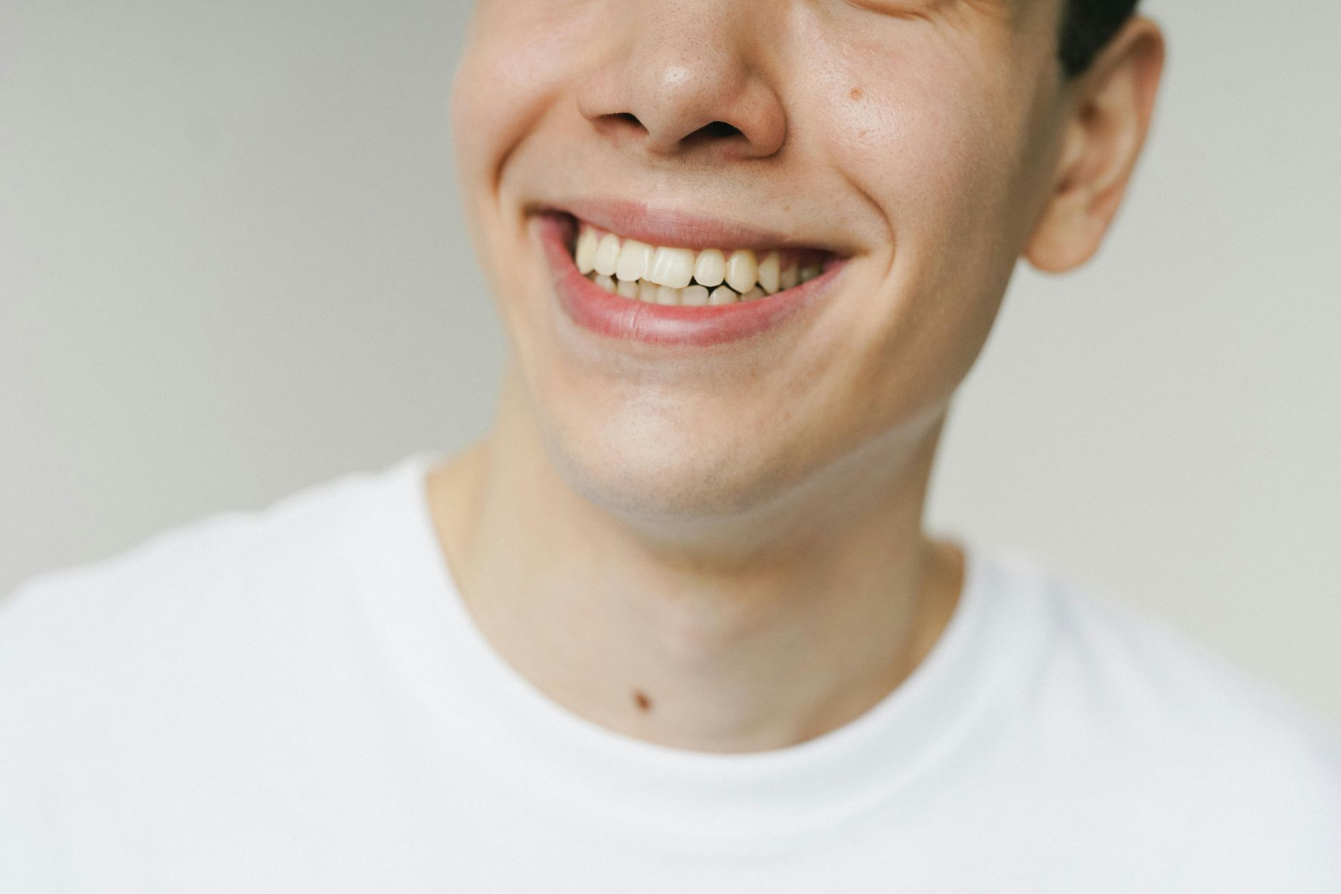 Man smiling, showing teeth, wearing a white t-shirt, against a light gray background.