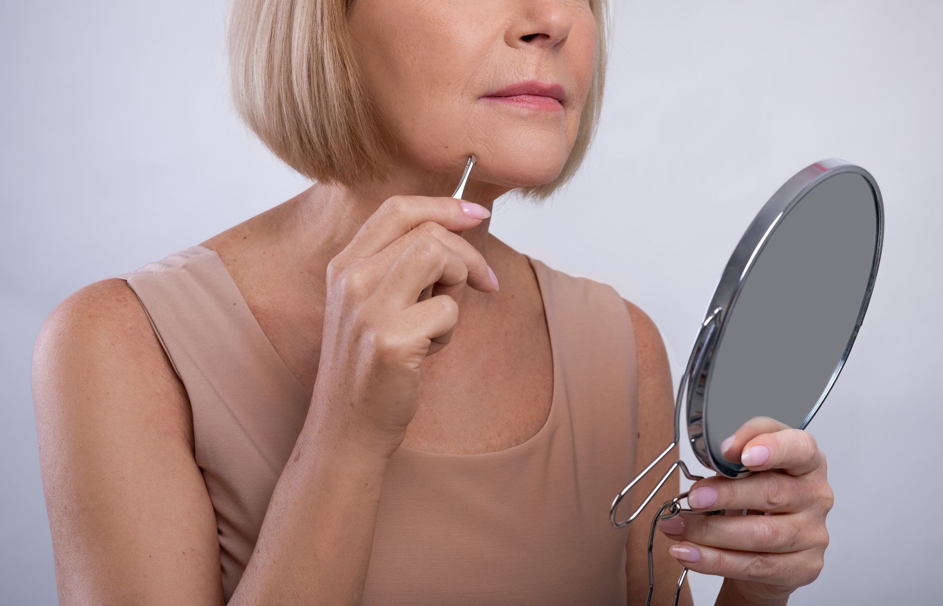Woman plucking chin hairs with tweezers, looking in a handheld mirror.